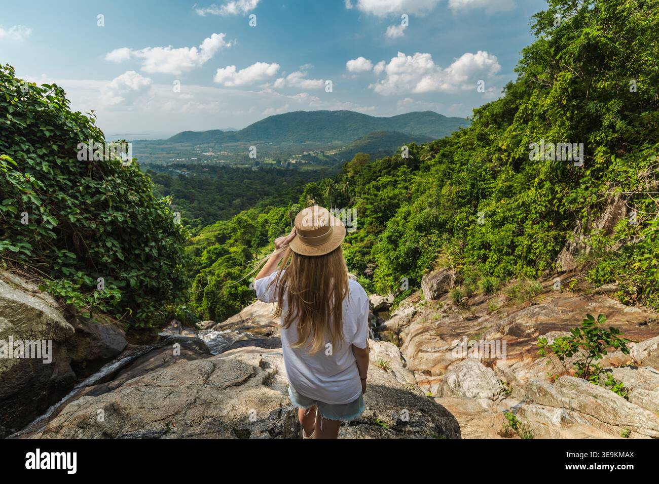 Eine junge Frau mit Strohhut auf einem felsigen Felsvorsprung mit Blick auf das üppige tropische Tal und die fernen Berge in der Nähe des Samui Wasserfalls in Thailand Stockfoto