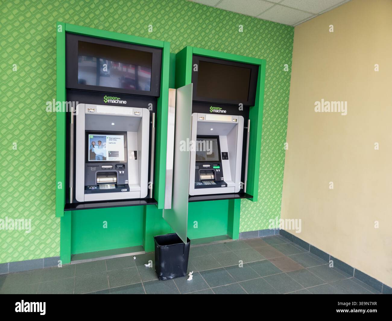 Zwei GreenMachine Geldautomaten, die nebeneinander in einem Bankbereich installiert wurden, einer aktiv und einer inaktiv, mit einem kleinen Mülleimer und verstreuten Papieren. - Smartphone-aufgenommenes Stockfoto