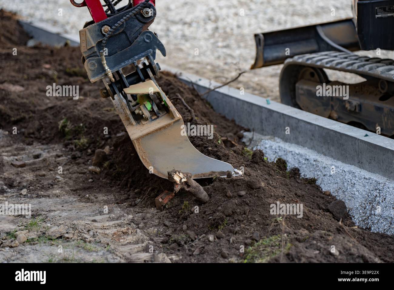 Hydraulischer Aufreißer für Bagger zieht Wurzeln durch dunklen Boden neben Betonkanne und Raupenmaschine für präzise Bodenreinigungsarbeiten Stockfoto