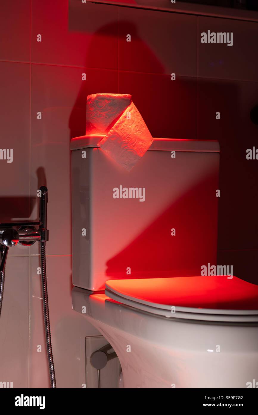 Rolle Toilettenpapier auf einer weißen Zisterne in dunkelrotem Licht. Konzeptionelle Visualisierung von Darmbewegungsproblemen, Verdauungsproblemen oder gastrointestinalen Symptomen. Fotografie Stockfoto