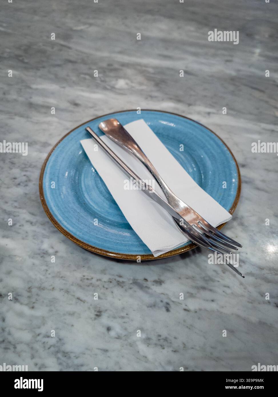 Ein minimalistischer Tisch mit einer hellblauen Keramikplatte, einer gefalteten Serviette und poliertem Besteck, das ordentlich angeordnet ist. Stockfoto