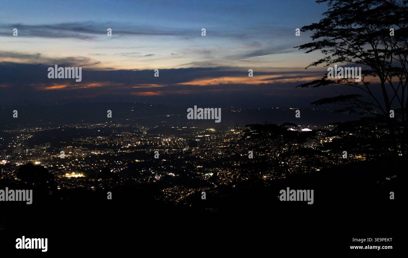 Panoramablick auf Bucaramanga, Kolumbien, beleuchtet bei Nacht unter einem tiefblauen Dämmerhimmel, eingerahmt von Baumsilhouetten und verblassenden Farben des Sonnenuntergangs Stockfoto