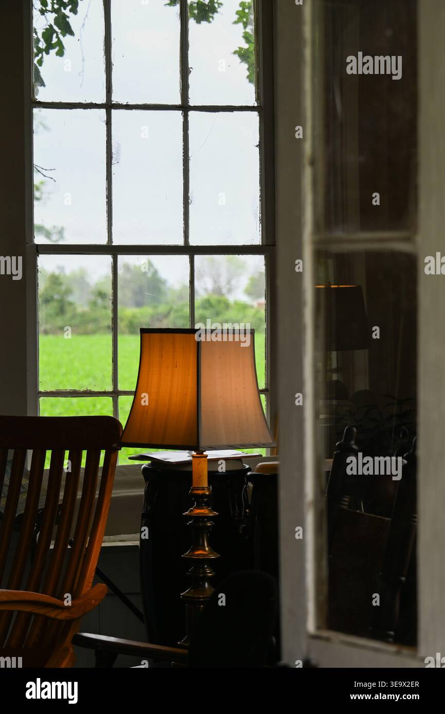 Kleine Hüttentür leicht offen, so dass ein Schaukelstuhl und eine Tischlampe von der Hinterleuchtung eines großen Fensters sichtbar sind Stockfoto
