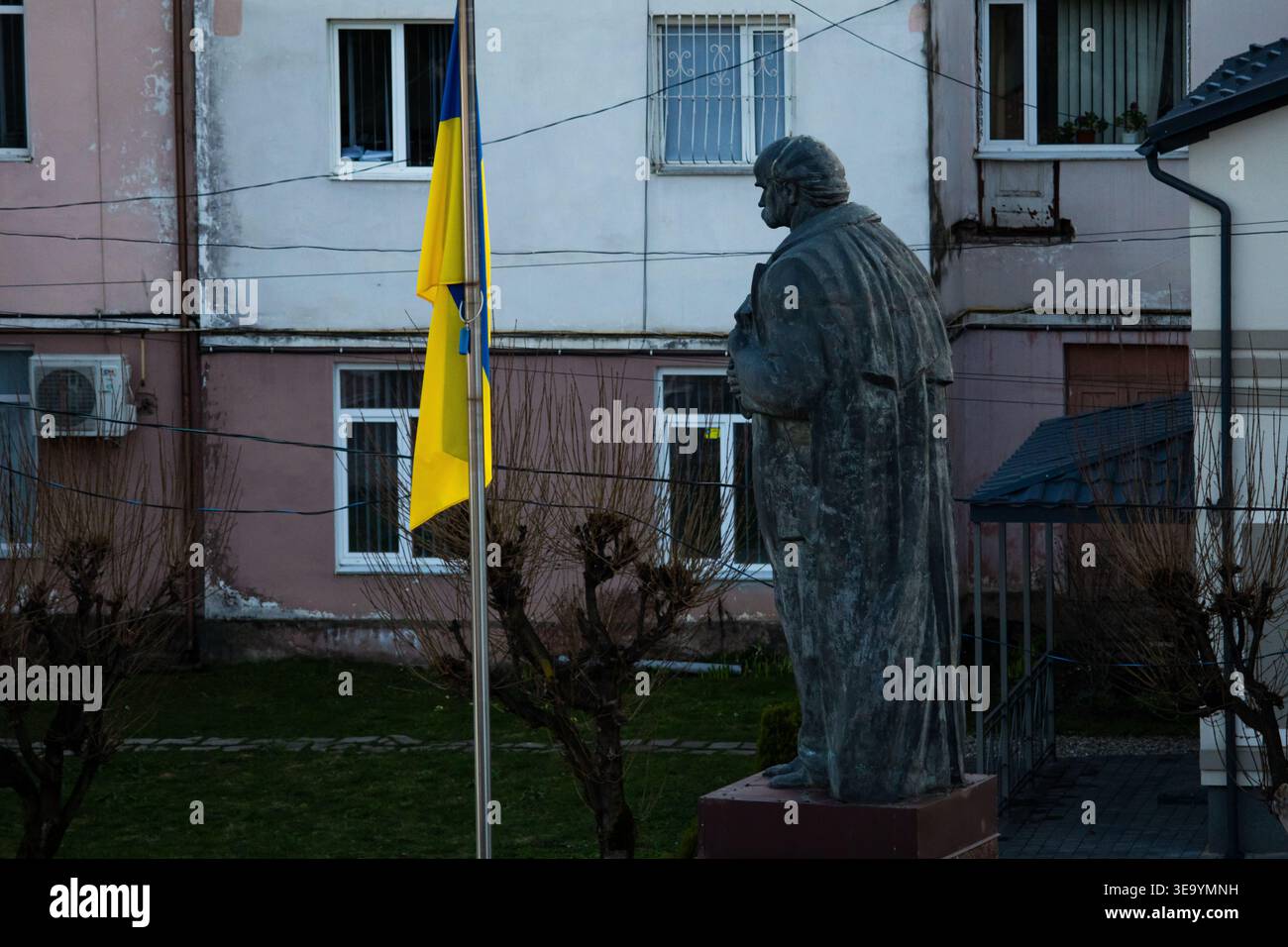 Denkmal einer gekleideten Figur neben einem verwitterten Gebäude mit einer ukrainischen Flagge, die in einer sanften Brise zum Ausdruck bringt, die an den Stolz der Bürger erinnert Stockfoto