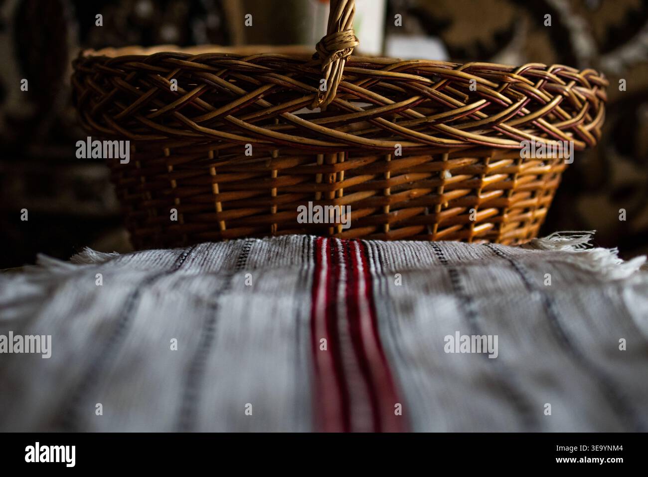 Der Korb sitzt über einem gefransten handgewebten Tuch mit einem kräftigen roten Streifen. Warme Töne und sanfte Schatten schaffen eine gemütliche rustikale Atmosphäre Stockfoto