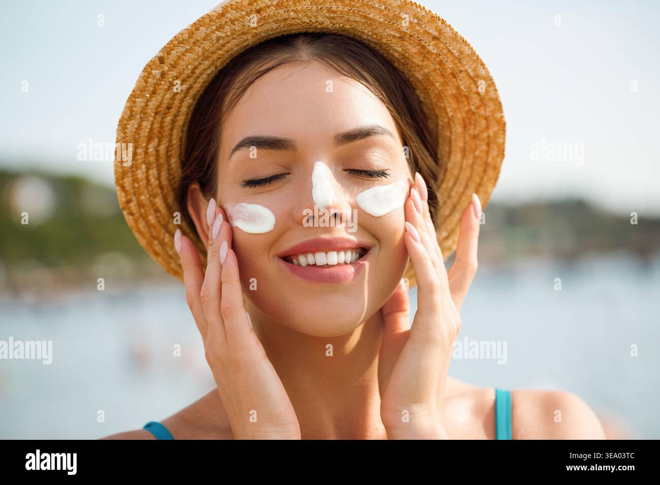 Frau, die das Gesicht mit beiden Händen berührt, mit Sonnencreme auf den Wangen. Eine junge Frau lächelt mit geschlossenen Augen. Weibliches Model genießt Hautpflegeroutine. Sonnenschutz Stockfoto