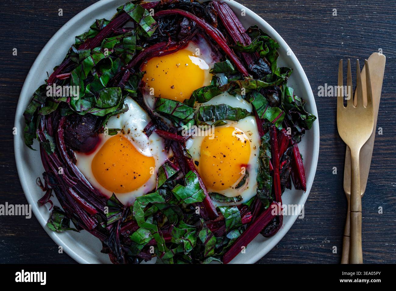 Ein nahrhaftes Frühstück: Drei Spiegeleier auf gebratenem Rüben-Grün und Stielen auf einem weißen Teller mit goldenem Besteck. Proteinreiche vegetarische Keto-Diät Stockfoto
