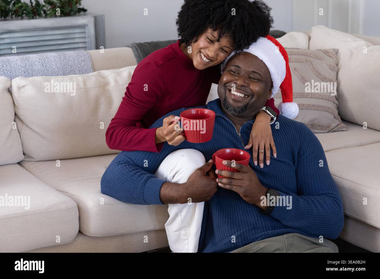 Afroamerikanisches Paar sitzt auf beigefarbenem Sofa, Mann mit Weihnachtsmütze und roten Tassen. Gemütlich, festlich, warm, Zuneigung, zusammen, Lächeln, zu Hause Stockfoto