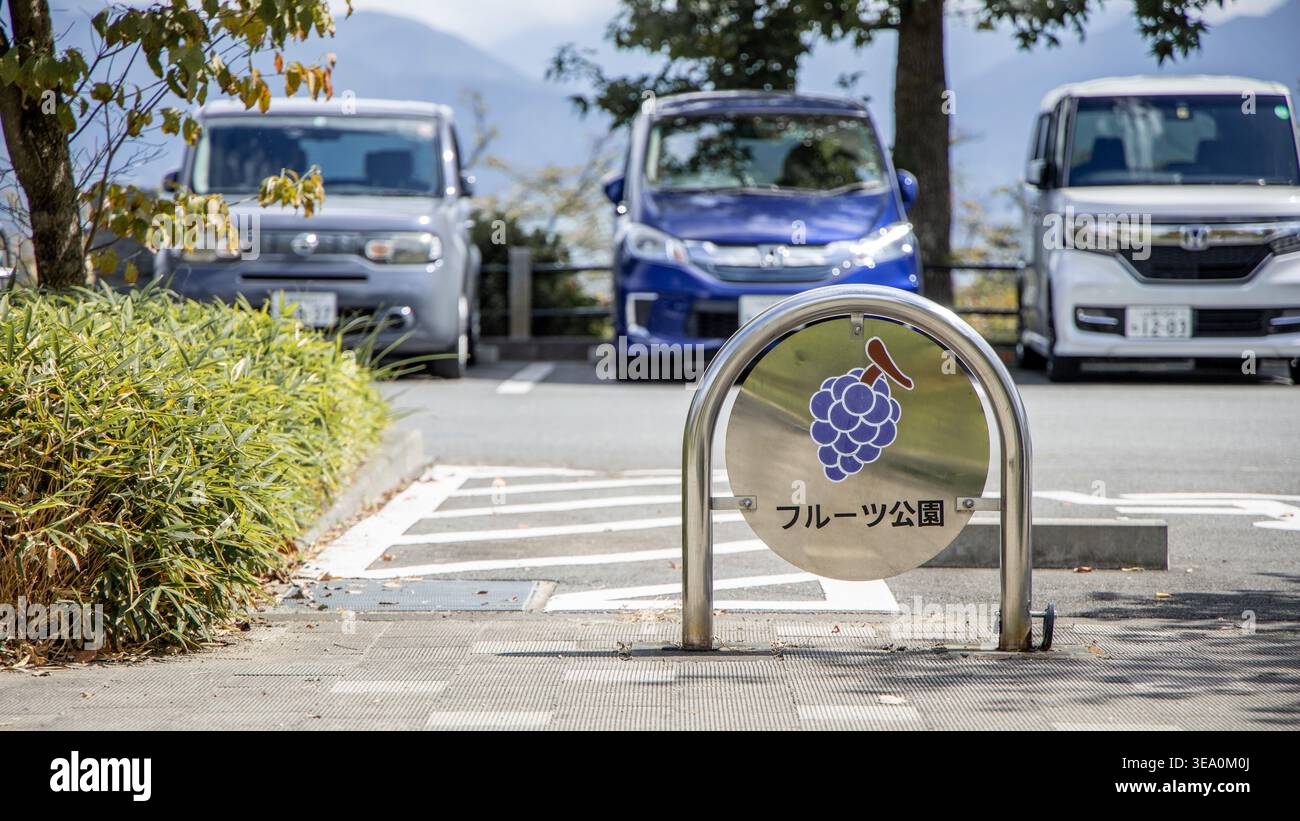 Fahrradständer und japanische Textschilder in einem Obstpark in Yamanashi, Japan, mit geparkten Fahrzeugen im Hintergrund. Stockfoto