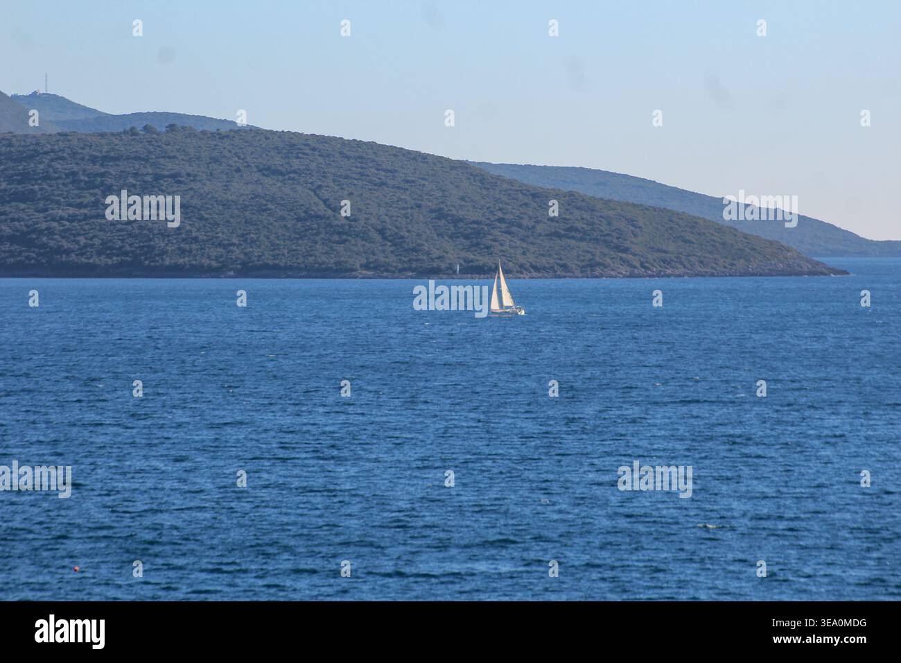 Kleines Segelboot gleitet auf ruhigem blauem Meer mit bewaldeten Hügeln im Hintergrund, friedliche Küstenlandschaft. Stockfoto