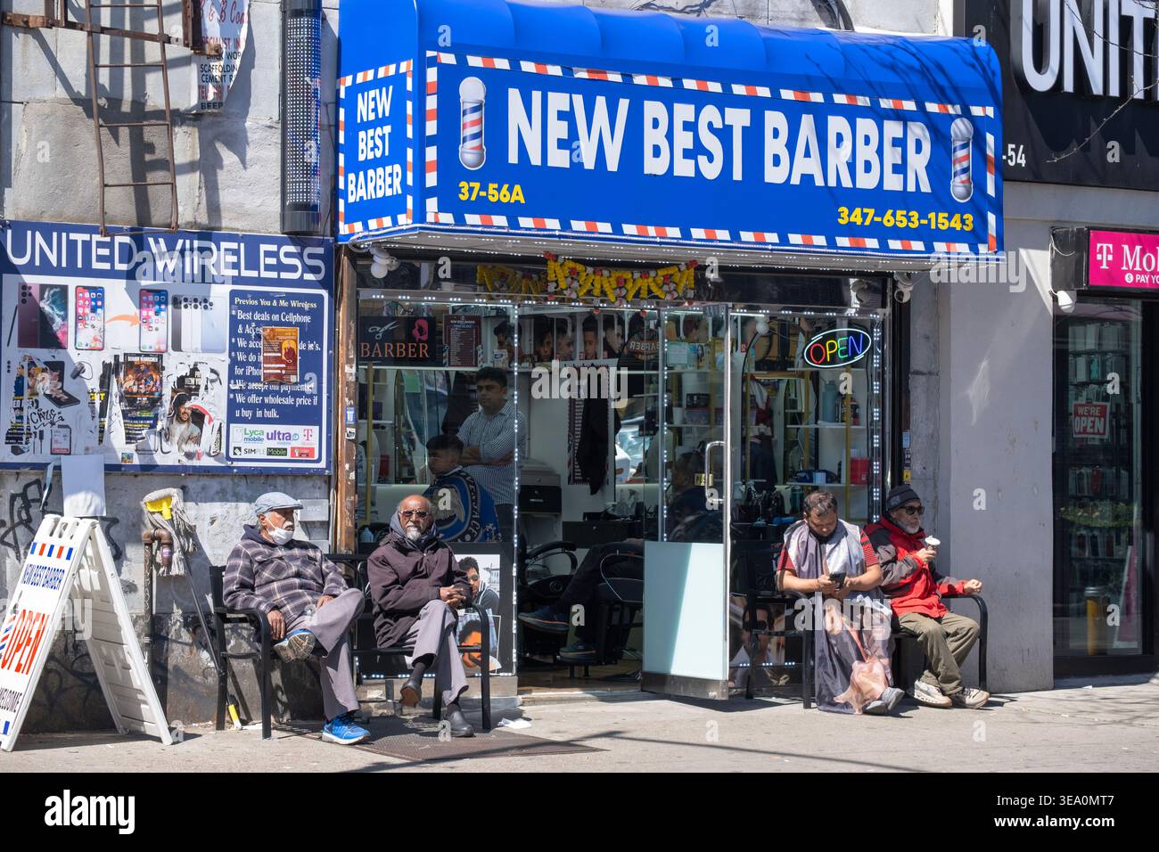 Straßenszene in Jackson Heights Queens, wo 4 nicht verbundene Männer vor dem New Best Friseur-Shop sitzen. An der 74th Street in Queens, New York. Stockfoto