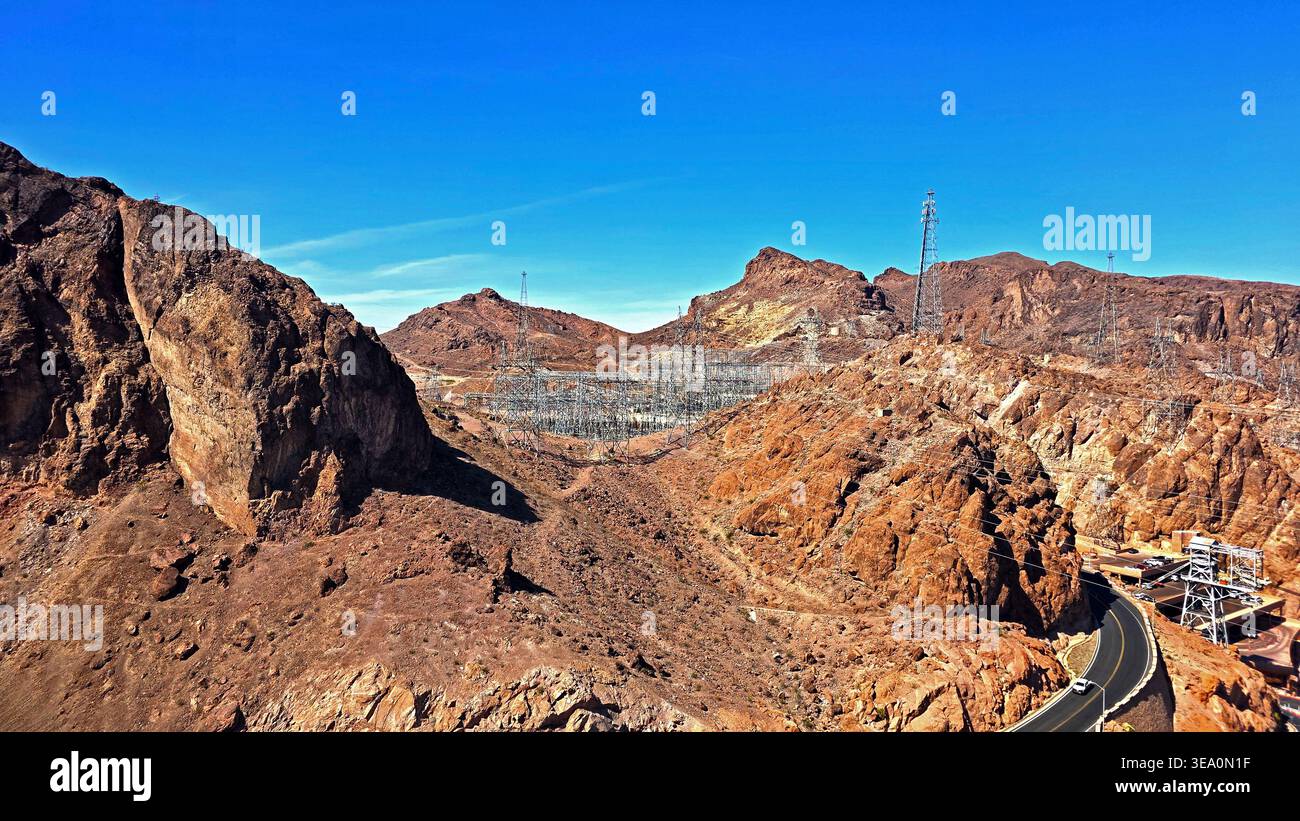 Das Stromnetz Der Wüste Nahe Dem Hoover-Staudamm. Felsige Wüstenlandschaft in der Nähe des Hoover Dam mit Übertragungstürmen, elektrischer Infrastruktur, kurviger Straße und klarem Wasser Stockfoto