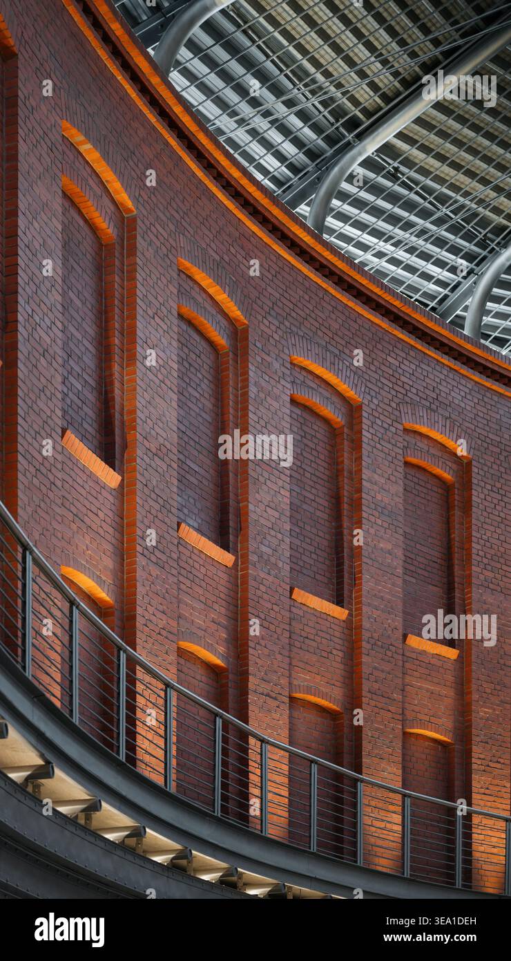 Gemauerte Mauer mit Bogennischen unter Stahldach. Eine geschwungene rote Backsteinfassade mit blinden Bogenfenstern und modernen Industriedetails. Stockfoto