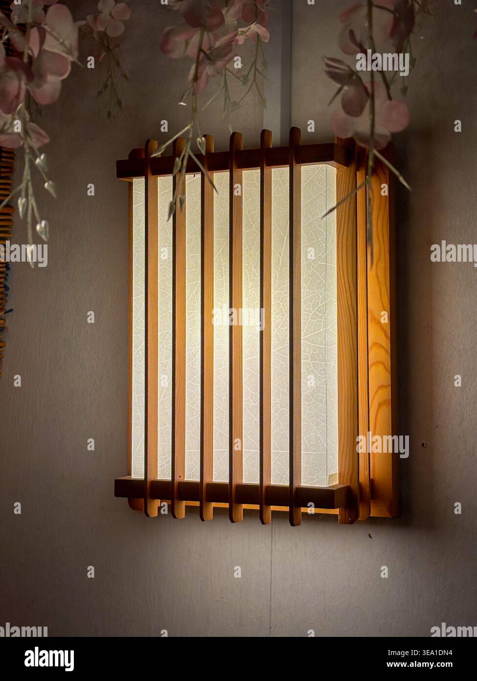 Holzlaterne hängt an einer strukturierten Wand. Weiches Licht filtert durch mattierte Glasplatten. Duftende rosa Blüten umrahmen die Laterne. Traditionelles Design A Stockfoto