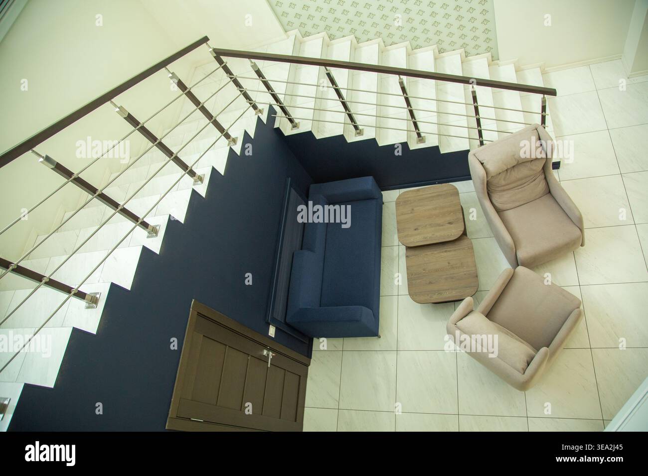 Hochwinkelaufnahme einer modernen Inneneinrichtung mit blauem Sofa, zwei beigefarbenen Sesseln und einem Holztisch neben einer weißen Treppe mit dunklem W Stockfoto