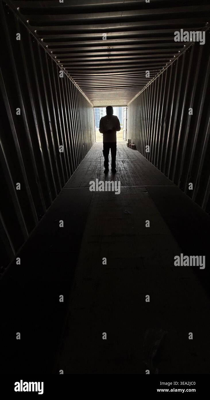 Eine Silhouette einer Person, die am Ende eines dunklen industriellen Containers steht, von hellem Tageslicht beleuchtet, was ein dramatisches und stimmungsvolles Ansehen erzeugt - Smartphone-aufgenommenes Stockfoto