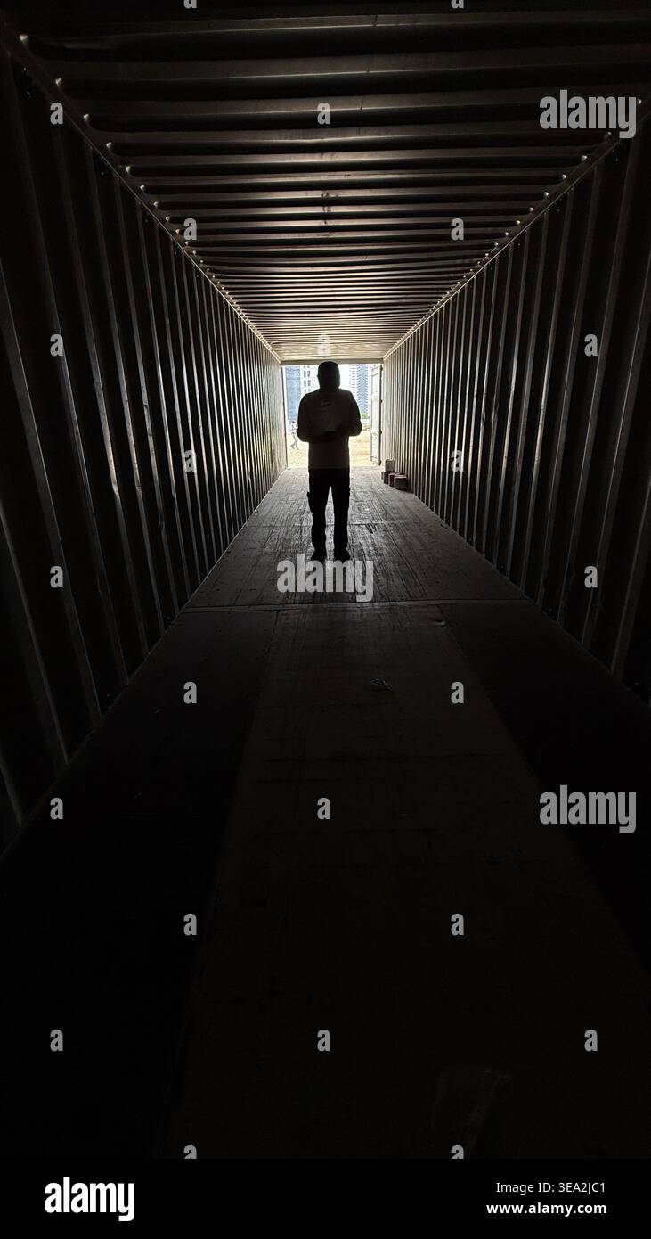 Eine Silhouette einer Person, die am Ende eines dunklen industriellen Containers steht, von hellem Tageslicht beleuchtet, was ein dramatisches und stimmungsvolles Ansehen erzeugt - Smartphone-aufgenommenes Stockfoto