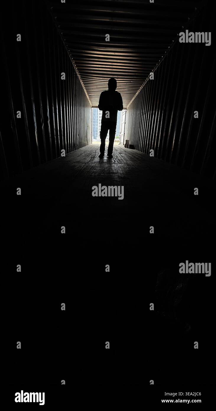 Eine Silhouette einer Person, die am Ende eines dunklen industriellen Containers steht, von hellem Tageslicht beleuchtet, was ein dramatisches und stimmungsvolles Ansehen erzeugt - Smartphone-aufgenommenes Stockfoto