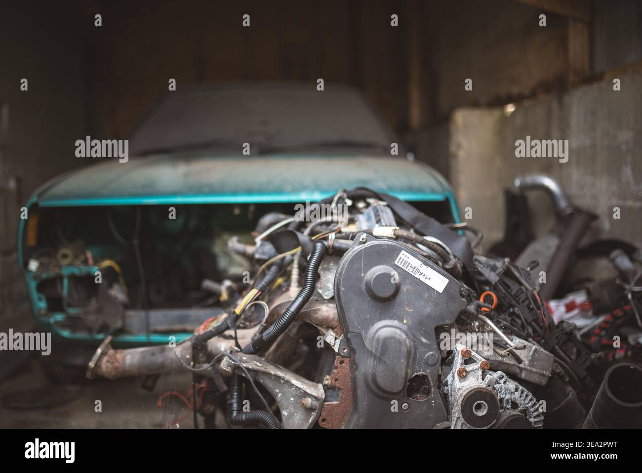 Ein altes, blaues Auto sitzt zerlegt in einer schwach beleuchteten Garage, mit einem Motor im Vordergrund. Stockfoto