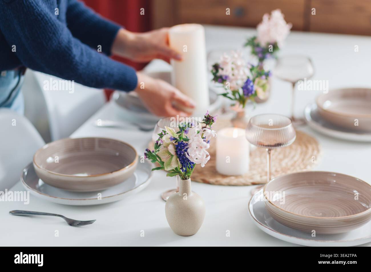 Frau dekoriert Esstisch mit Frühlingsblumen in Vasen und Kerzen zu Hause. Nahaufnahme von Geschirr, Besteck für vier. Stockfoto