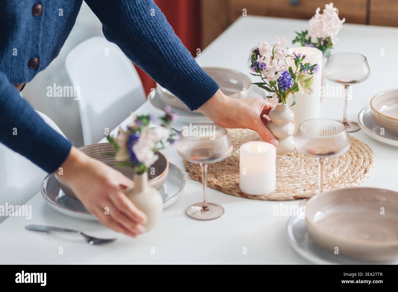 Frau dekoriert Esstisch mit Frühlingsblumen in Vasen und Kerzen zu Hause. Nahaufnahme von Geschirr, Besteck für vier. Stockfoto