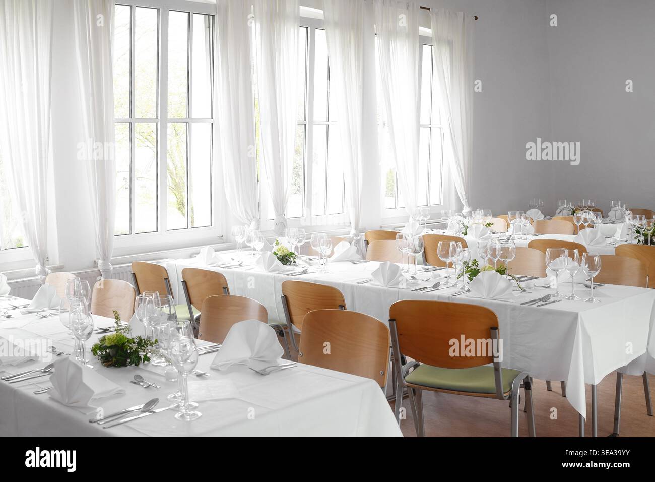 Festliche Tischdekoration mit weißen Tischdecken, gefalteten Servietten, Gläsern und Besteck in einem modernen Tagungsraum mit großen Fenstern und hellen Vorhängen Stockfoto
