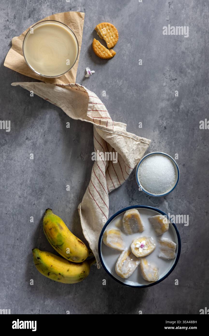 Eine Nahaufnahme einer Banane in Kokosnussmilch, serviert in einer Schüssel mit einem Glas Bananen-Smoothie und Reifen Bananen, einem traditionellen thailändischen Dessert. Draufsicht. Flach l Stockfoto