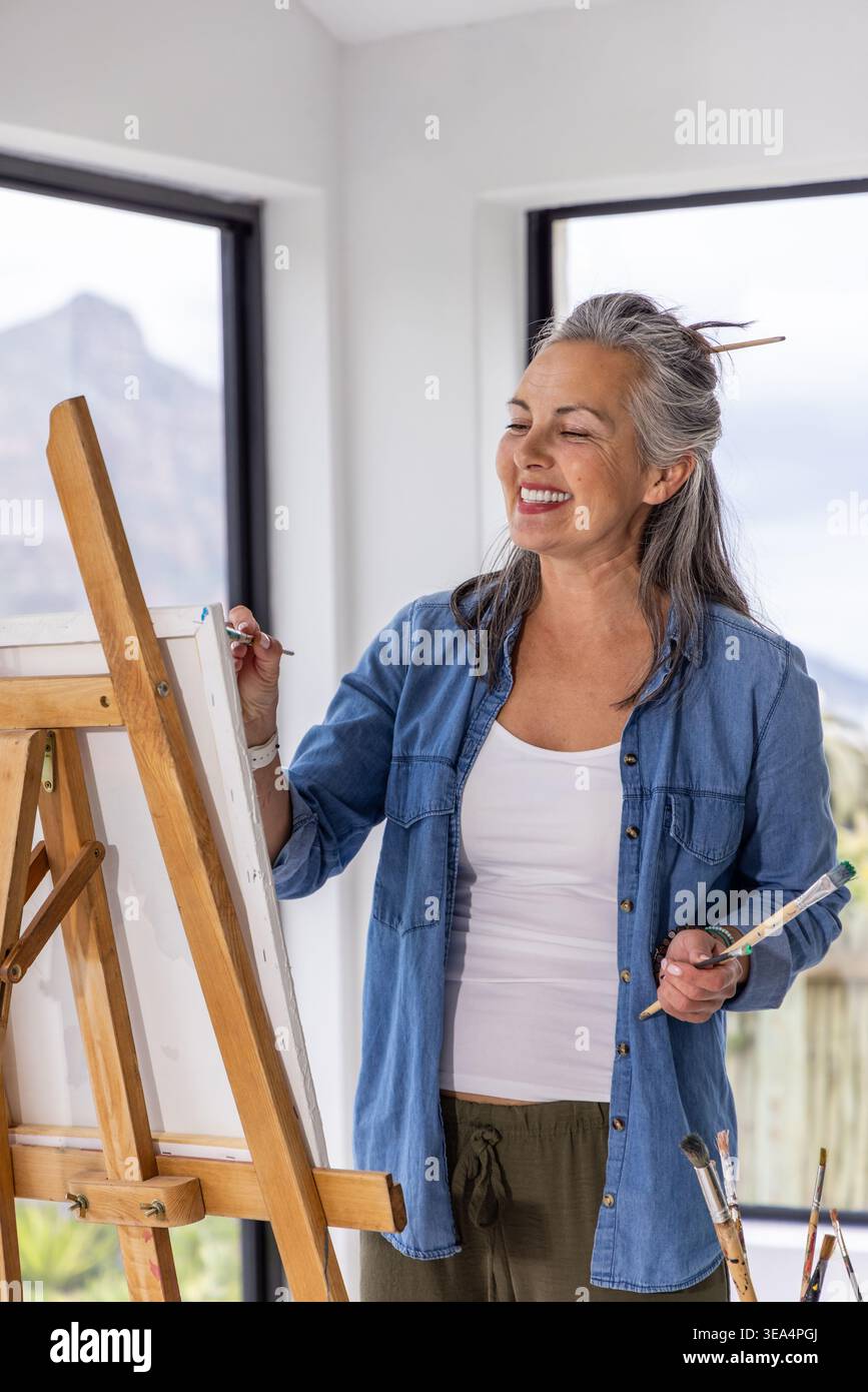 Eine ältere Künstlerin, die an einer hölzernen Staffelei auf Leinwand in einem hellen Atelier mit Pinseln steht Stockfoto