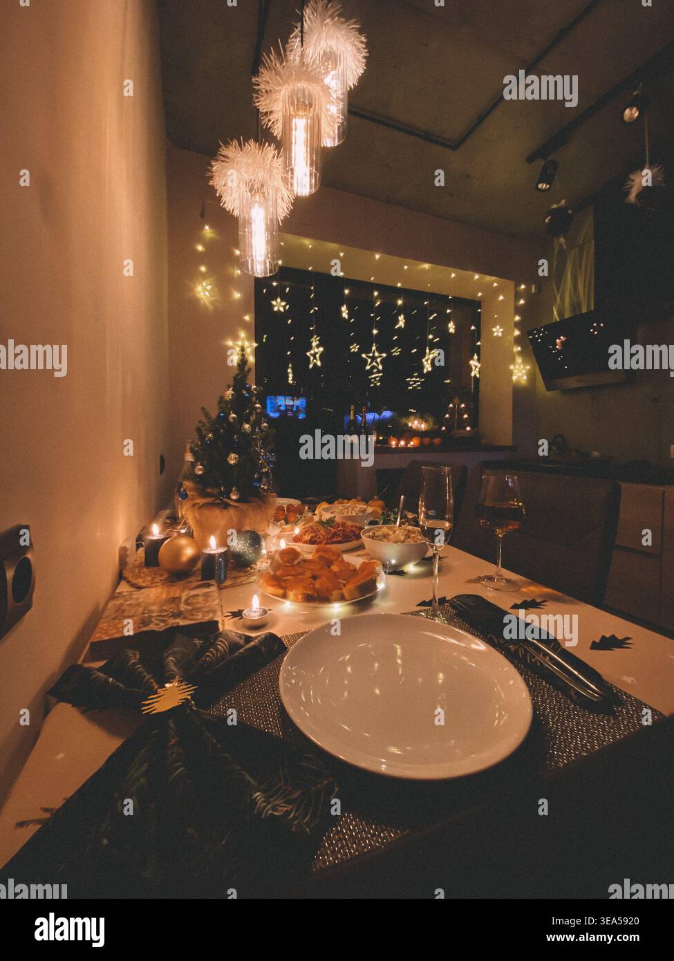 Gemütlicher Weihnachts-Esstisch mit festlicher Dekoration, Kerzen, Essen und Wein in einem warmen, beleuchteten Wohnraum bei Nacht. - Smartphone-aufgenommenes Stockfoto