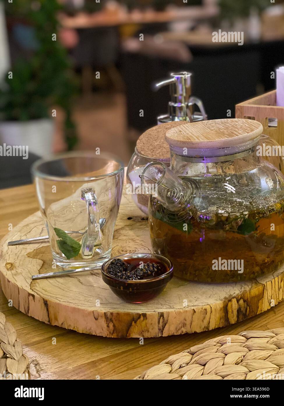 Glas-Teekanne mit Kräutertee, Honig und Minzblättern auf einem Holztablett in einem gemütlichen Café-Interieur, warme und entspannende Atmosphäre. - Smartphone-aufgenommenes Stockfoto