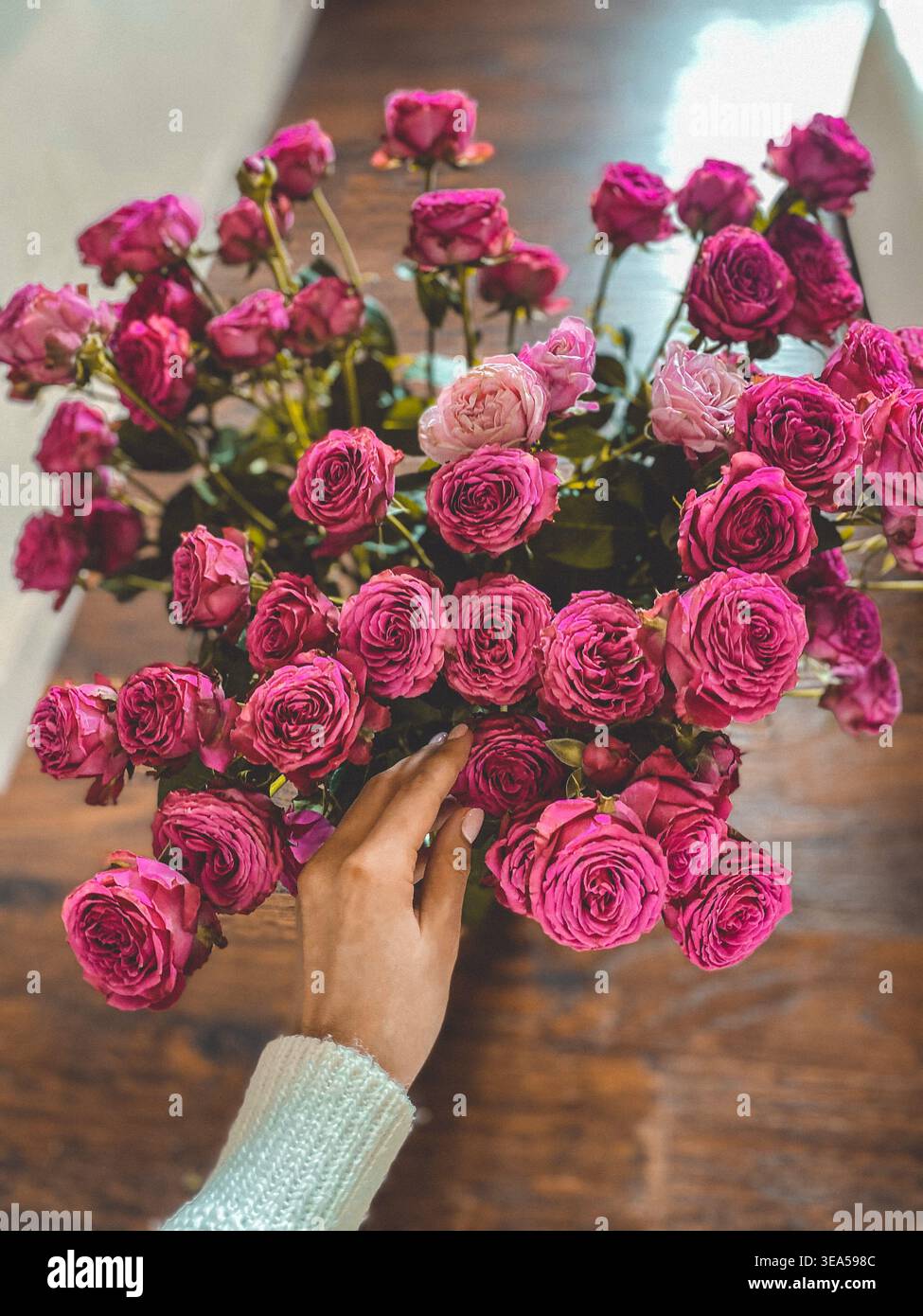 Blumenstrauß aus rosa Rosen mit einer weiblichen Hand, die Blumen auf einem Holztisch berührt, sanftes natürliches Licht und romantische Atmosphäre. - Smartphone-aufgenommenes Stockfoto