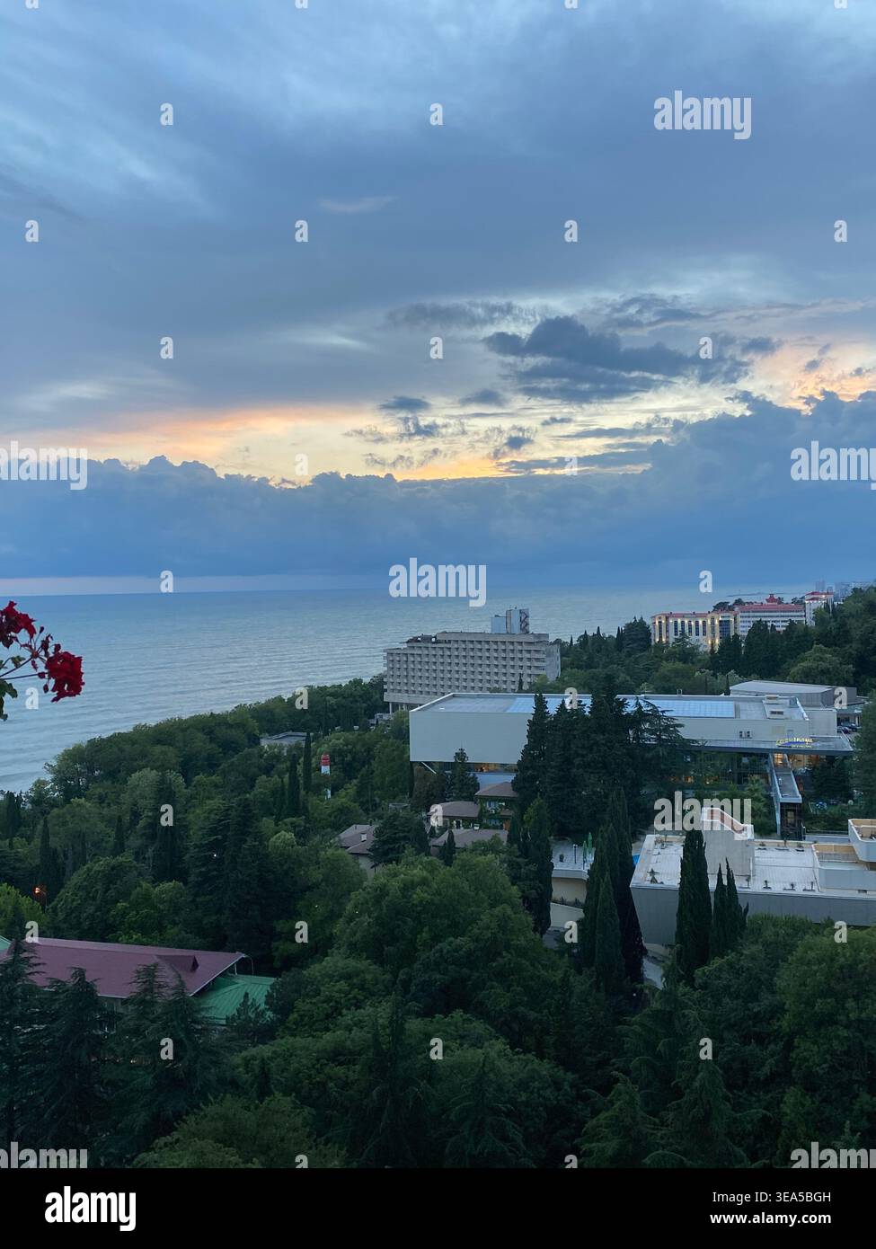 Blick auf die Küstenstadt mit Meer und dramatisch bewölktem Sonnenuntergang über grüner Landschaft und Gebäuden - Smartphone-aufgenommenes Stockfoto