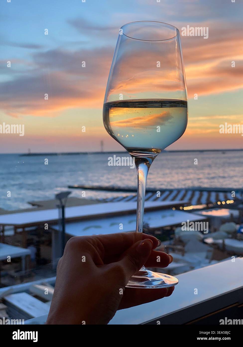 Hand hält ein Glas Weißwein vor einem wunderschönen Sonnenuntergang am Meer, mit weichem pastellfarbenem Himmel im Glas und ruhigem Ozean im Hintergrund - Smartphone-aufgenommenes Stockfoto