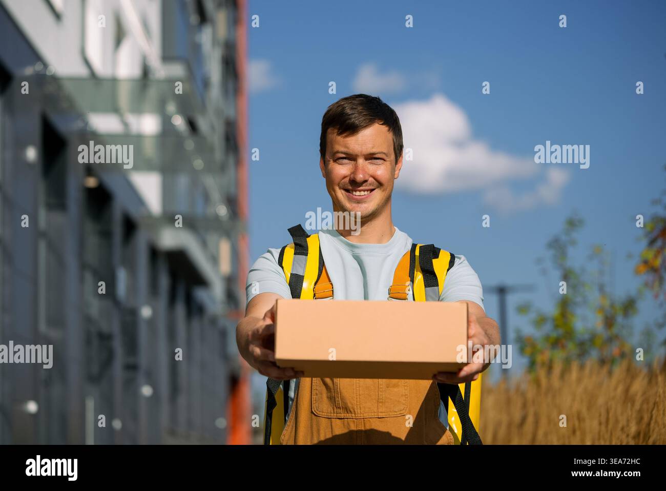 Ein lächelnder junger Kurier, der ein Paket liefert, einen isolierten Lieferrucksack und Overall trägt, draußen vor einem modernen Stadtgebäude steht und einen ausgezeichneten Versandservice bietet Stockfoto