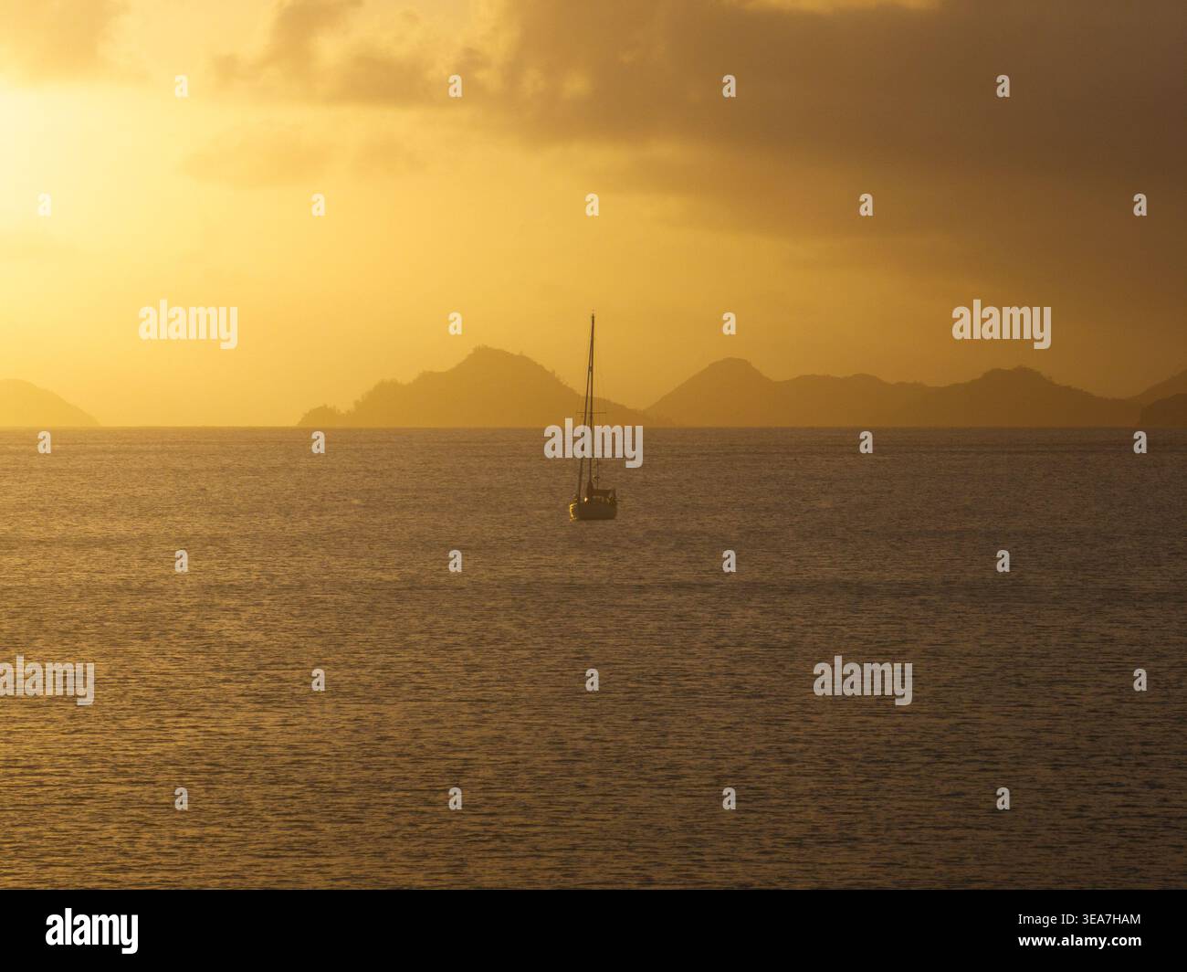 Aus der Vogelperspektive eines einsamen Segelboots auf dem ruhigen Wasser von Anse A La Mouche während eines goldenen Sonnenuntergangs mit Inselsilhouetten am Horizont der Seychellen. Stockfoto