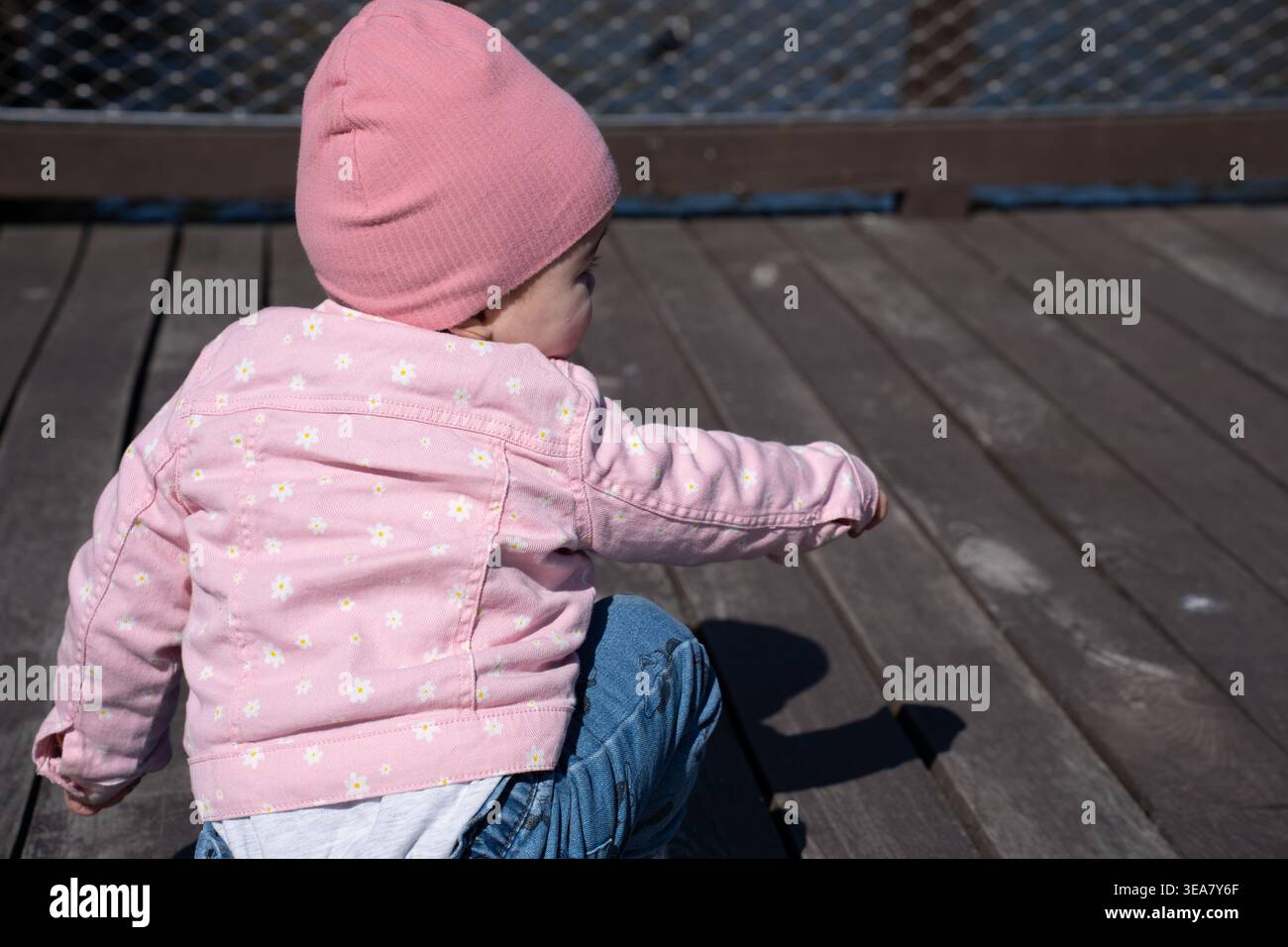 Rückansicht eines Kleinkindes, das auf einem sonnendurchfluteten Holzsteg sitzt, mit rosa Beanie und Blumenjacke nach außen reicht; starker Schatten, verwitterte Dielen Stockfoto