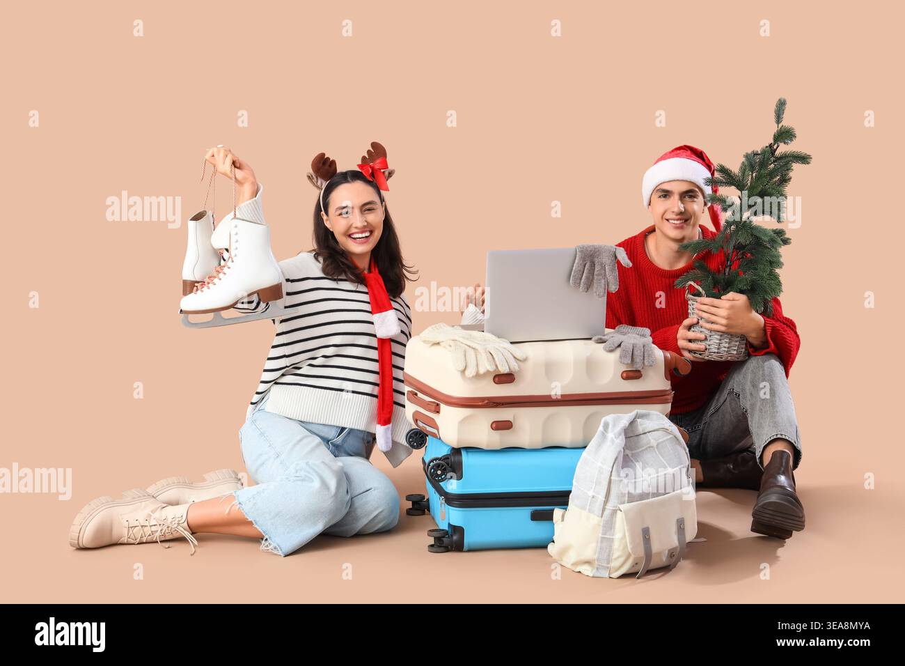 Nettes junges Paar mit Laptop, Koffern und kleinem Weihnachtsbaum auf beigefarbenem Hintergrund. Winterurlaubskonzept Stockfoto