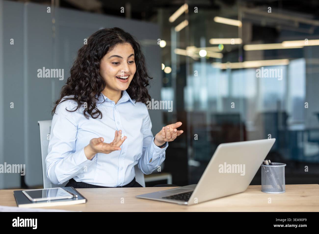 Junge indische Geschäftsfrau, die ein virtuelles Meeting auf einem Laptop in einem modernen Büro hat, während sie mit Gesten an einem Schreibtisch kommuniziert und ihre Arbeit diskutiert Stockfoto