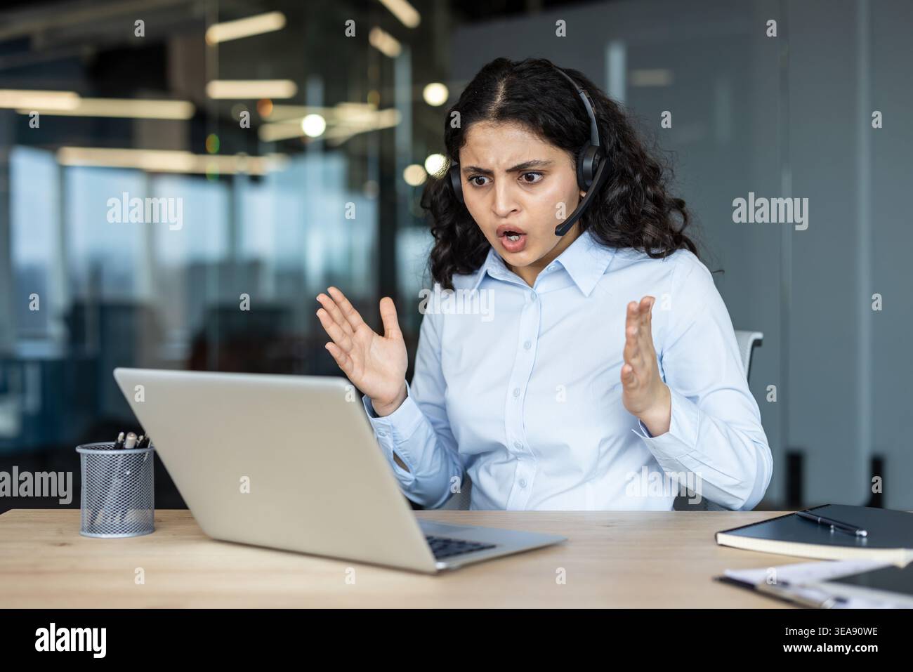 Gestresster junger Agent mit Headset und Uniform, der Wut durch Gesten mit den Händen ausdrückt, während er an einem Laptop in einer modernen Kundendienstumgebung arbeitet Stockfoto