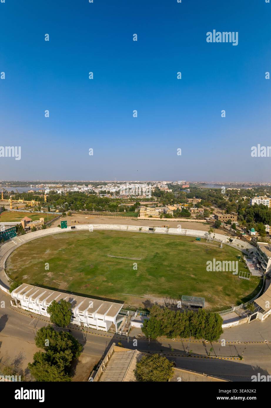 Blick aus der Vogelperspektive auf das Niaz Stadium mit seinem grünen Cricketfeld und weißen Sitzplätzen, umgeben von urbaner Landschaft und Bäumen unter einem klaren blauen Himmel in Hy Stockfoto