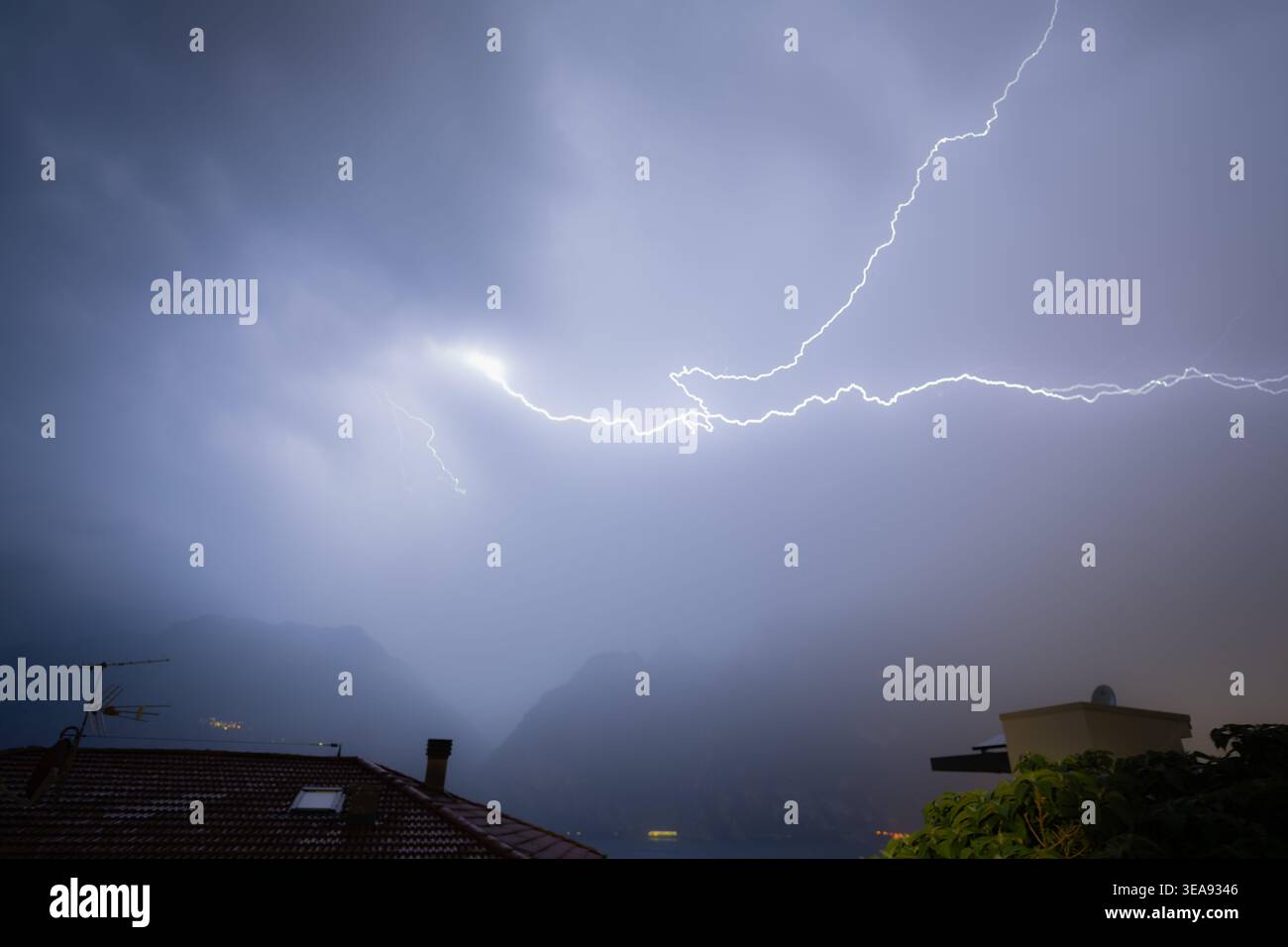 Blitze erleuchten ein dramatisches Gewitter über dem gardasee mit Bergen und Dächern darunter Stockfoto