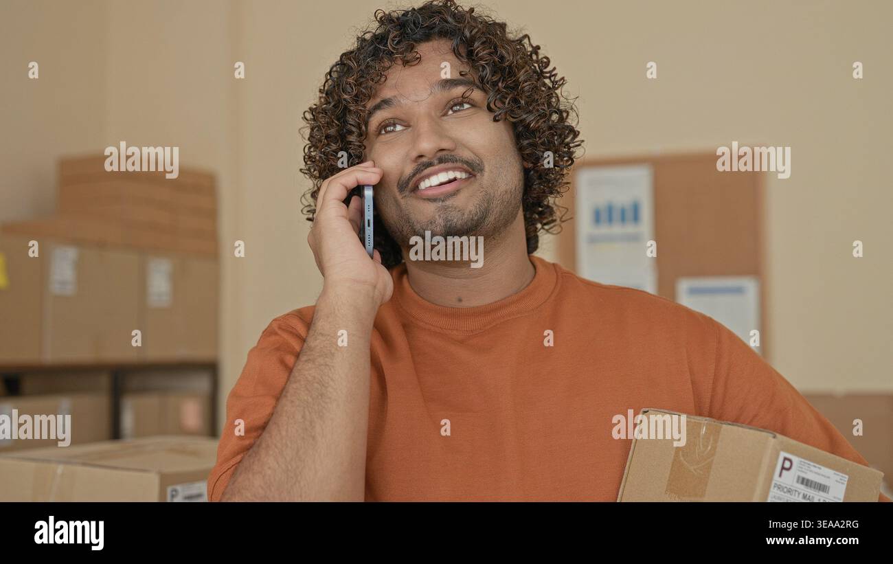 Mann mit Pappkarton und Smartphone, Hand an Ohr beim Lächeln und Reden, umgeben von Paketen im Gebäude; Optimismus für kleine Unternehmen. Stockfoto