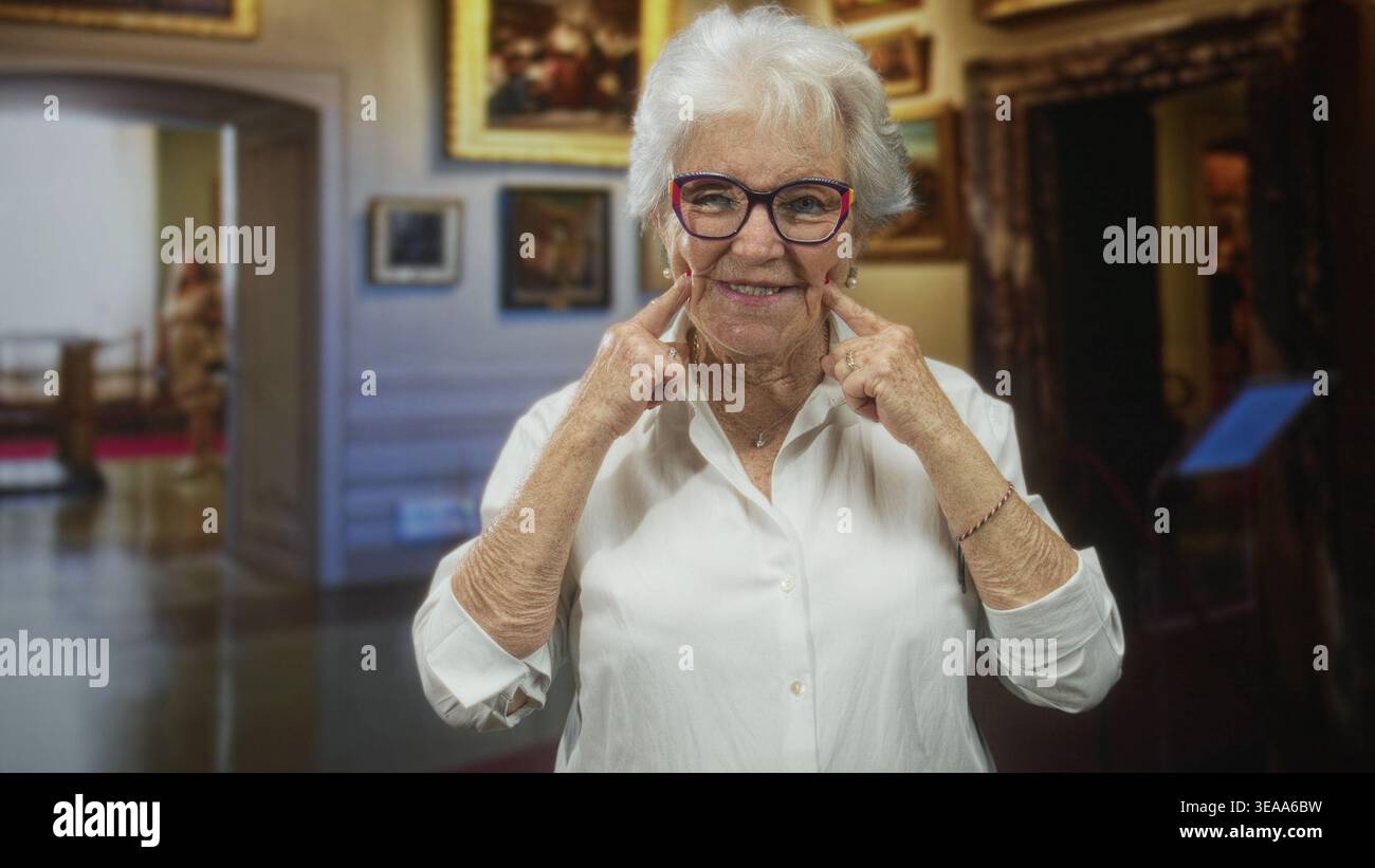 Ältere Frau mit Brille zeigt Zeigefinger auf die Wangen, während sie in einem Museumsgebäude zwischen gerahmten Gemälden lächelt; Freude Nostalgie. Stockfoto