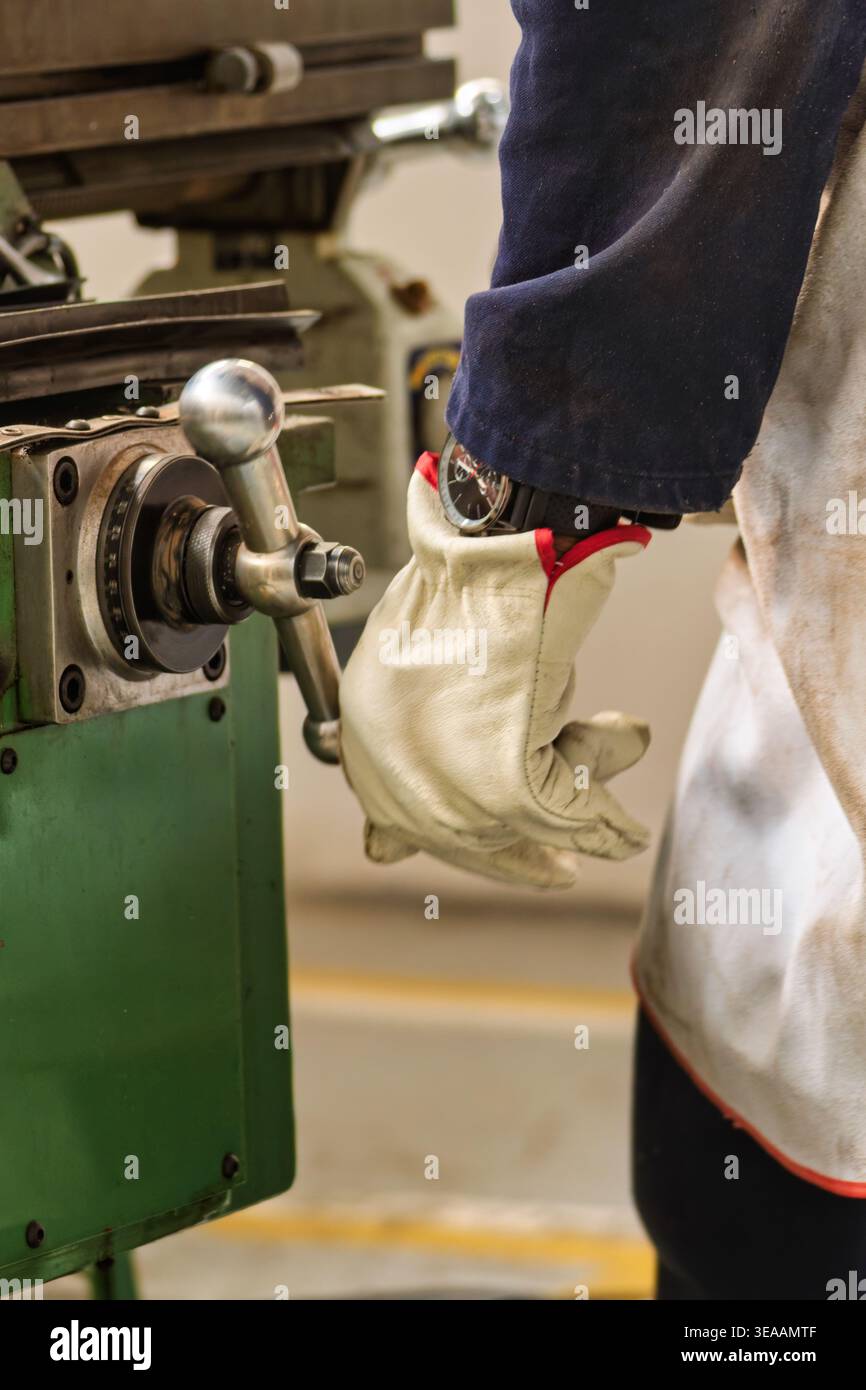 Ein Maschinenführer mit Schutzhandschuh und Armbanduhr bedient das Metallhandrad einer industriellen Dreh- oder Fräsmaschine in einer Werkstatt. Stockfoto