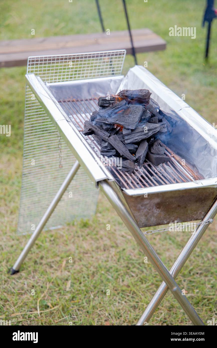 Holzkohleverbrennung auf einem tragbaren Edelstahl-Grill auf einem Outdoor-Campingplatz, glühende Glut sichtbar unter der Schwarzkohle. Klassische Sommerkochszene im Freien. Stockfoto