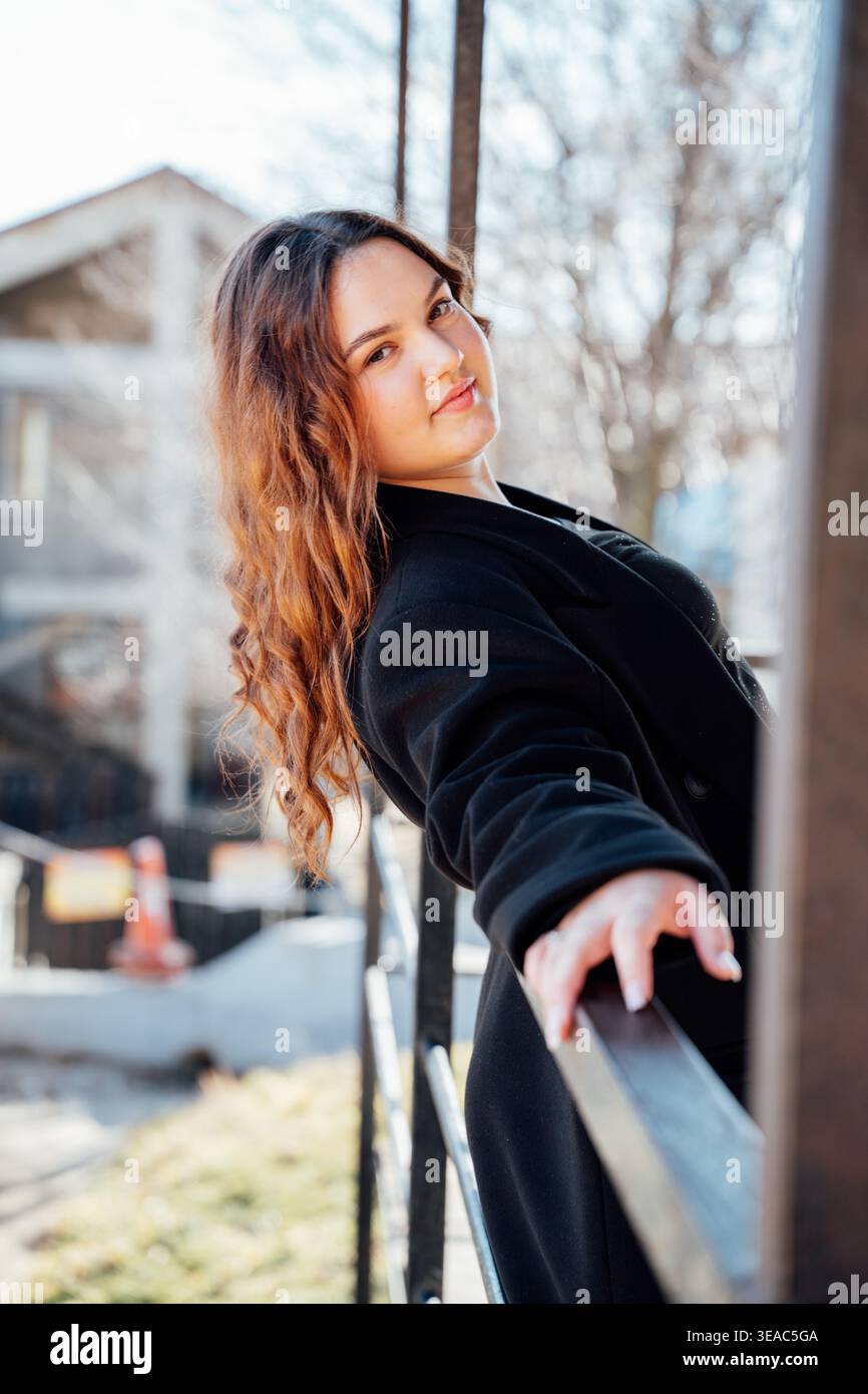 Junge kaukasische Frau mit langen, welligen Haaren in schwarzem Mantel, gestützt auf Metallgeländer draußen in der Stadt und blickt bei Tageslicht in die Kamera. Hochformat, li Stockfoto