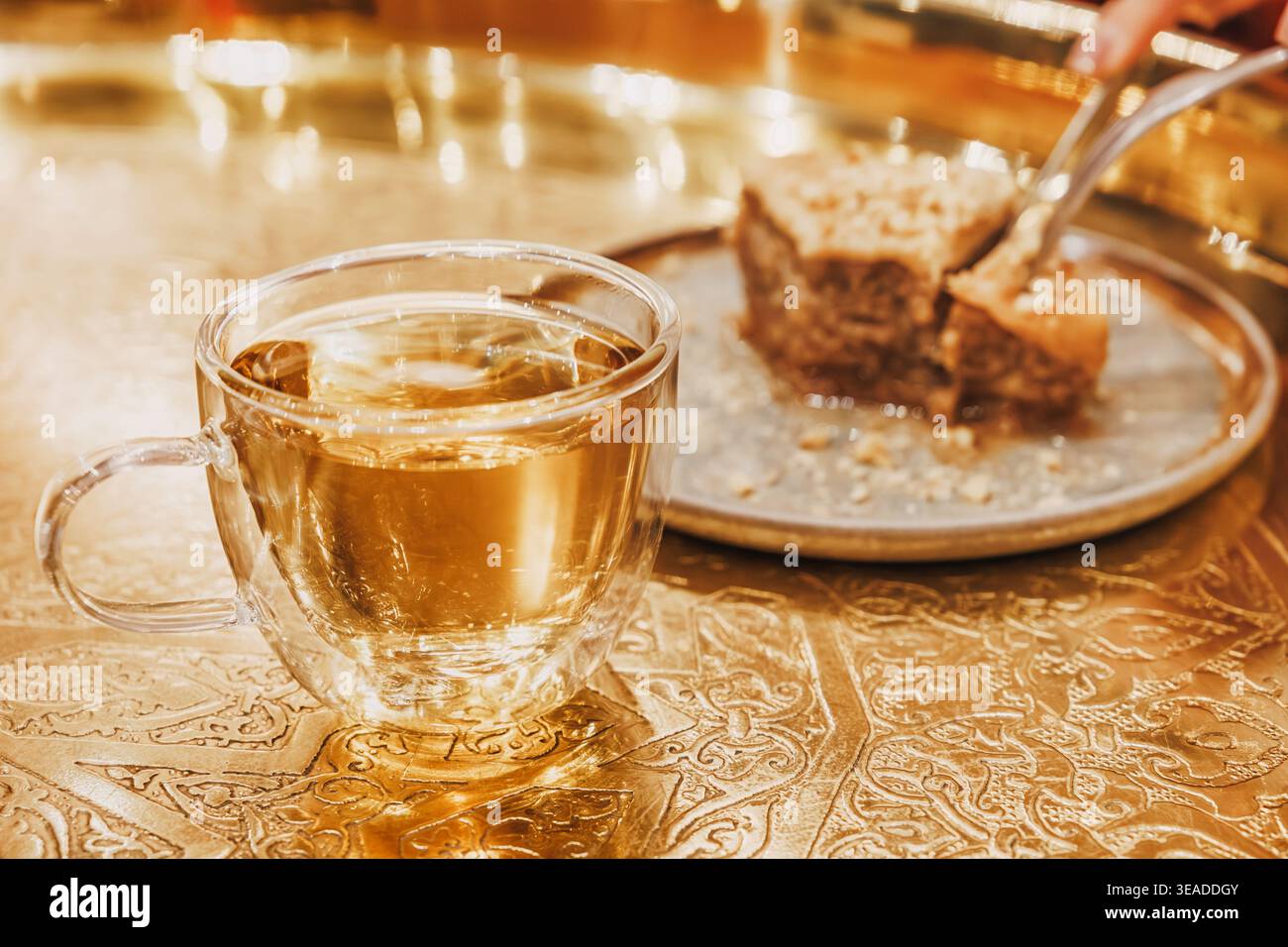 Tee in einer Glasschale und eine Person, die ein Stück Baklava von einem Teller auf einem kunstvollen goldenen Tablett nimmt. Traditionelles Dessert Stockfoto