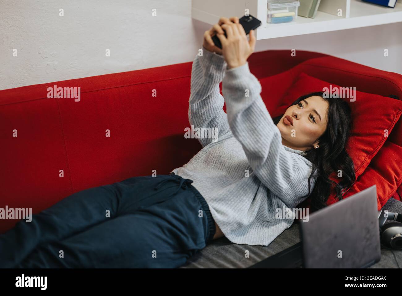 Studentin, die auf einer roten Couch liegt und ein Selfie mit Telefon neben einem Laptop macht, während sie studiert Stockfoto