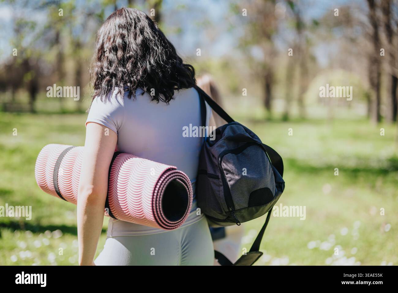 Frau mit Yogamatte und Sporttasche, die durch den sonnigen Park geht, bereit für Outdoor-Workout Stockfoto