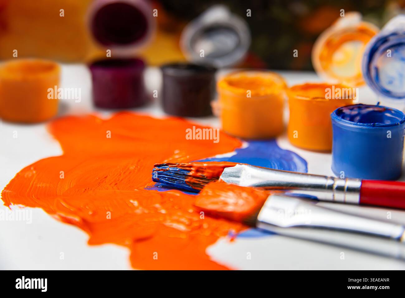Künstler mischen orange Farbe mit Pinseln auf einem Tisch, der von verschiedenen Farbbehältern umgeben ist, in einem gut beleuchteten Bereich während einer kreativen Aktivität. Stockfoto